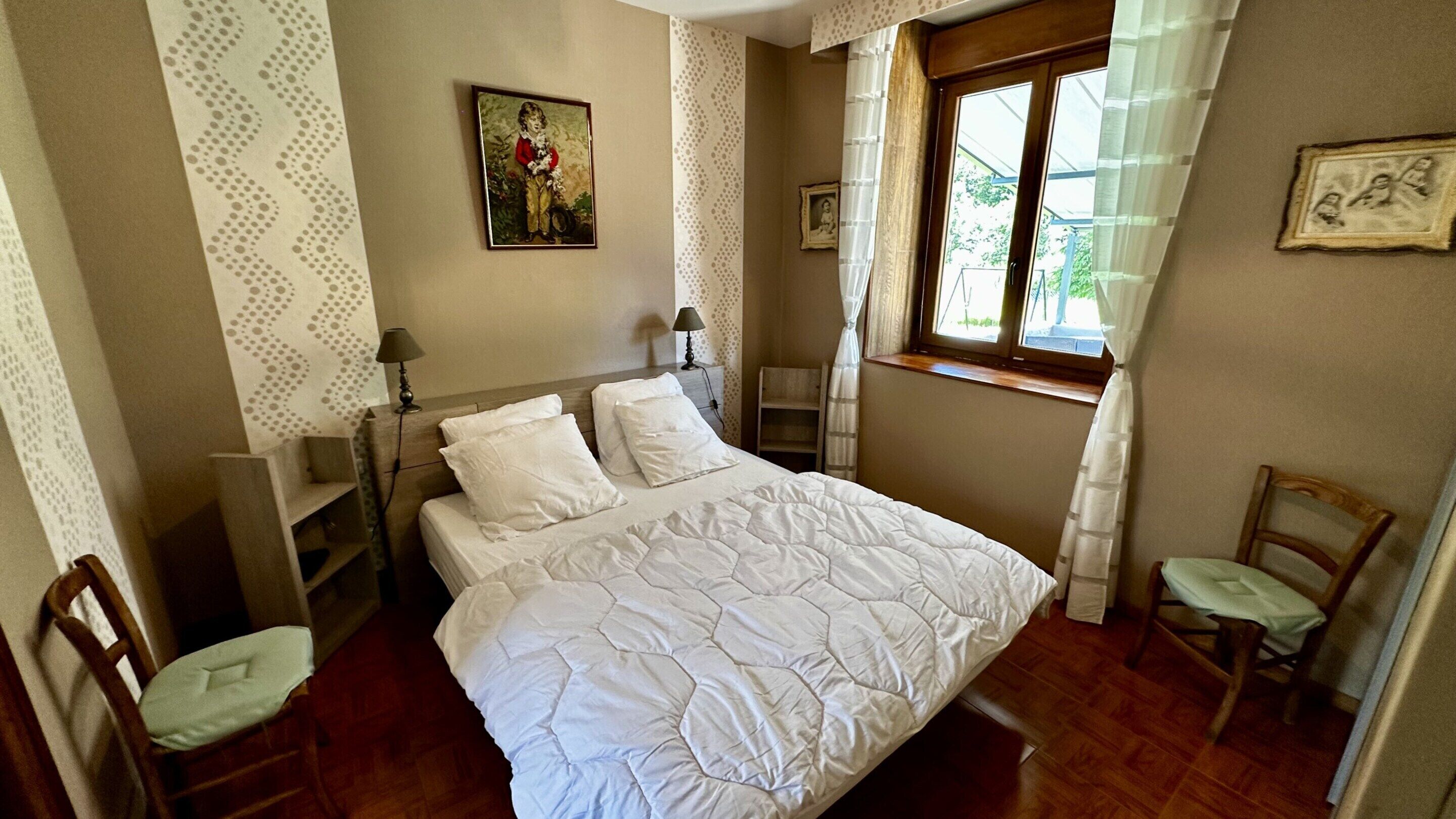 Ein ordentlich bezogenes Bett mit weißer Decke, ein Nachttisch aus Holz mit Lampe, ein Stuhl und ein Fenster mit Vorhängen.