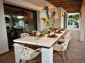 A dining area with a long table set for six, surrounded by white chairs with cut-out backrests. - Luxury villa with large private pool within walking distance of beach, bars & stores (Calpe)