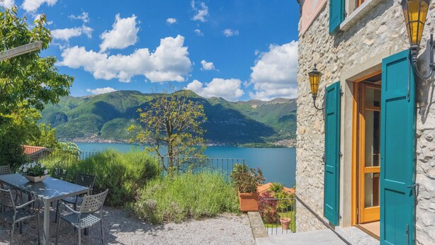 Un edificio de piedra con una terraza, una mesa y sillas, y vistas a un lago y montañas.