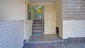 Una escalera conduce a una puerta con panel de vidrio, suelo de baldosas y una rejilla de ventilación en la pared.