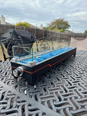 A table with a glass container holding blue pebbles, a metal grill, and a metal bench.