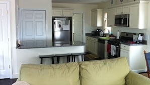Una cocina con armarios blancos, un refrigerador de acero inoxidable, un microondas y una estufa. En primer plano, un sofá color beige.
