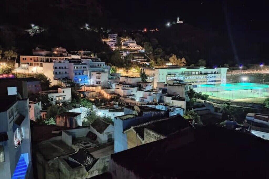 Une vue nocturne d’un village perché sur une colline, avec des bâtiments illuminés, un terrain de sport et une structure située au sommet d’une colline au loin.