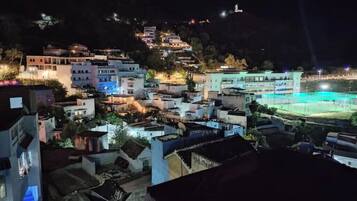 Une vue nocturne d’un village perché sur une colline, avec des bâtiments illuminés, un terrain de sport et une structure située au sommet d’une colline au loin.