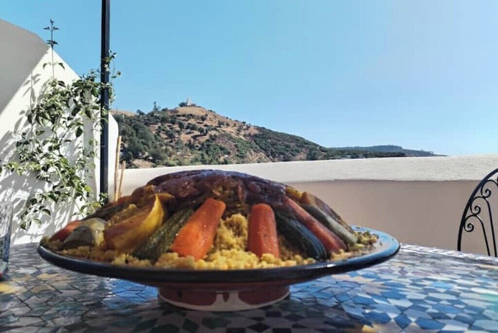 Una bandeja grande de verduras y cuscús sobre una mesa con suelo de baldosas, con un cerro y cielo despejado al fondo.