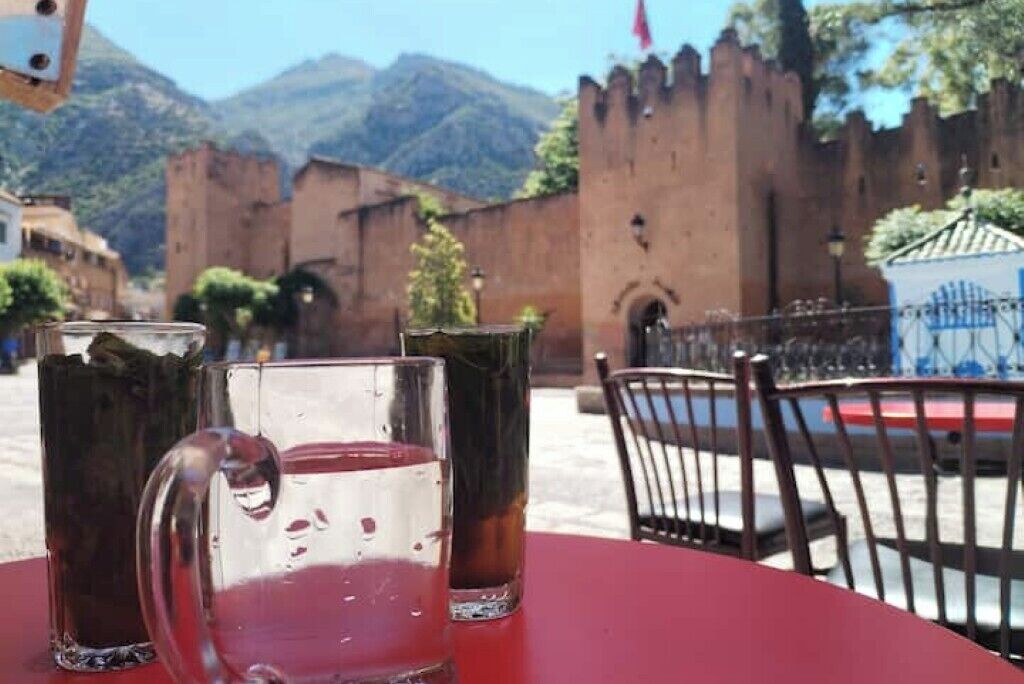 Ein Tisch mit zwei Gläsern und einer Karaffe vor einer historischen Gebäudeecke mit Tor und Bergen im Hintergrund.