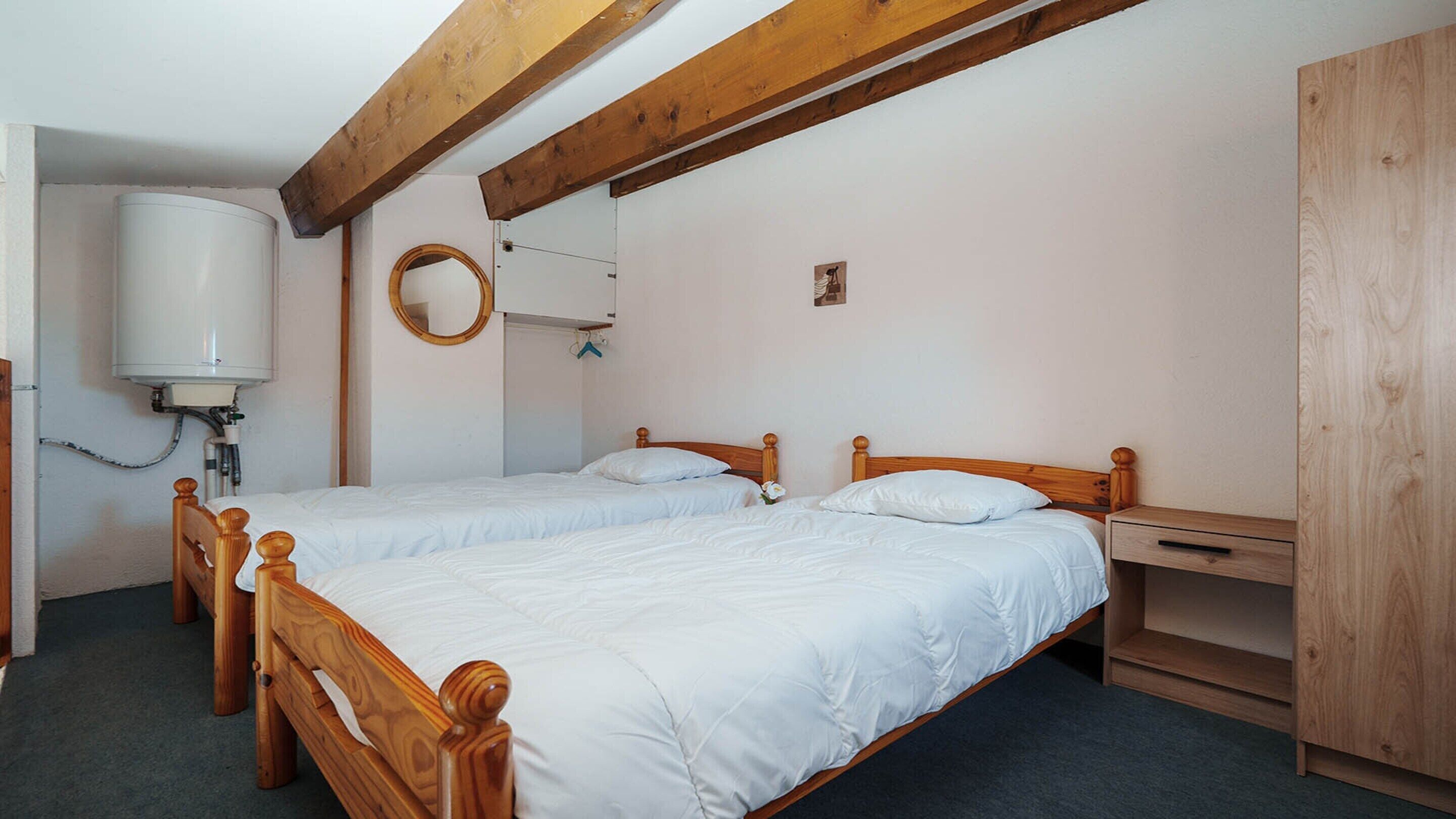 Une chambre avec deux lits en bois, une armoire en bois, une petite table de chevet, un miroir et un radiateur blanc.