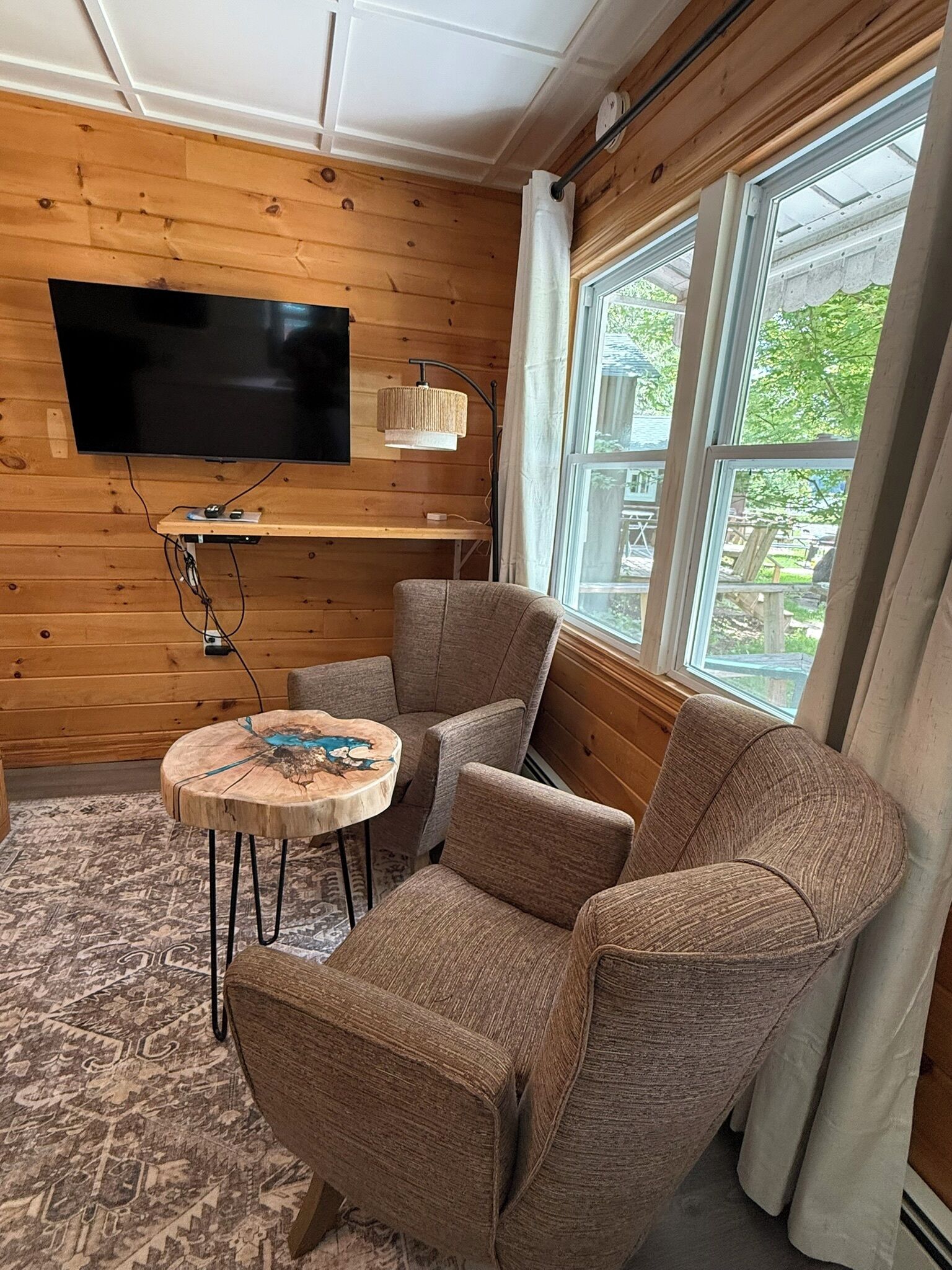 A cozy room with wooden walls, a flat-screen TV mounted on the wall, a round wooden coffee table, two upholstered armchairs, and a window with curtains.