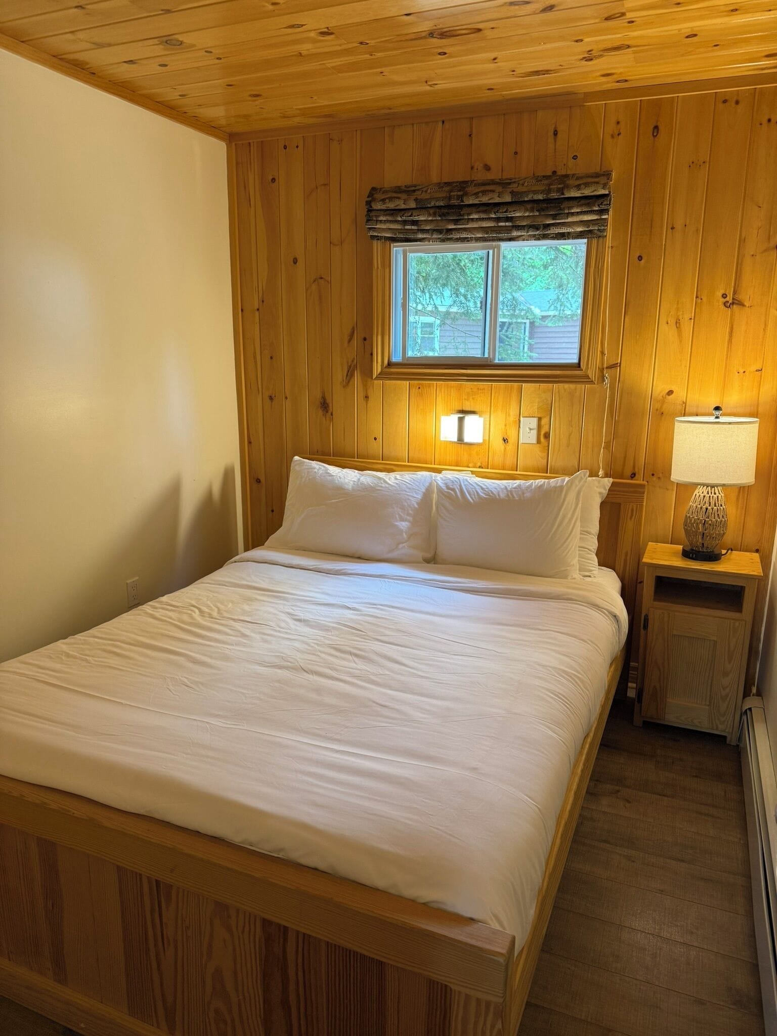 A wooden-paneled room with a bed, a window with a view, and a nightstand with a lamp.