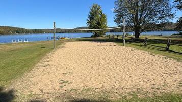 A sandy beach area with a volleyball net, trees, and a lake in the background.