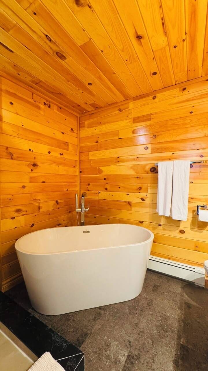 A wooden cabin interior with a large bathtub, wooden walls, and a small bathroom with a mirror and towel rack.