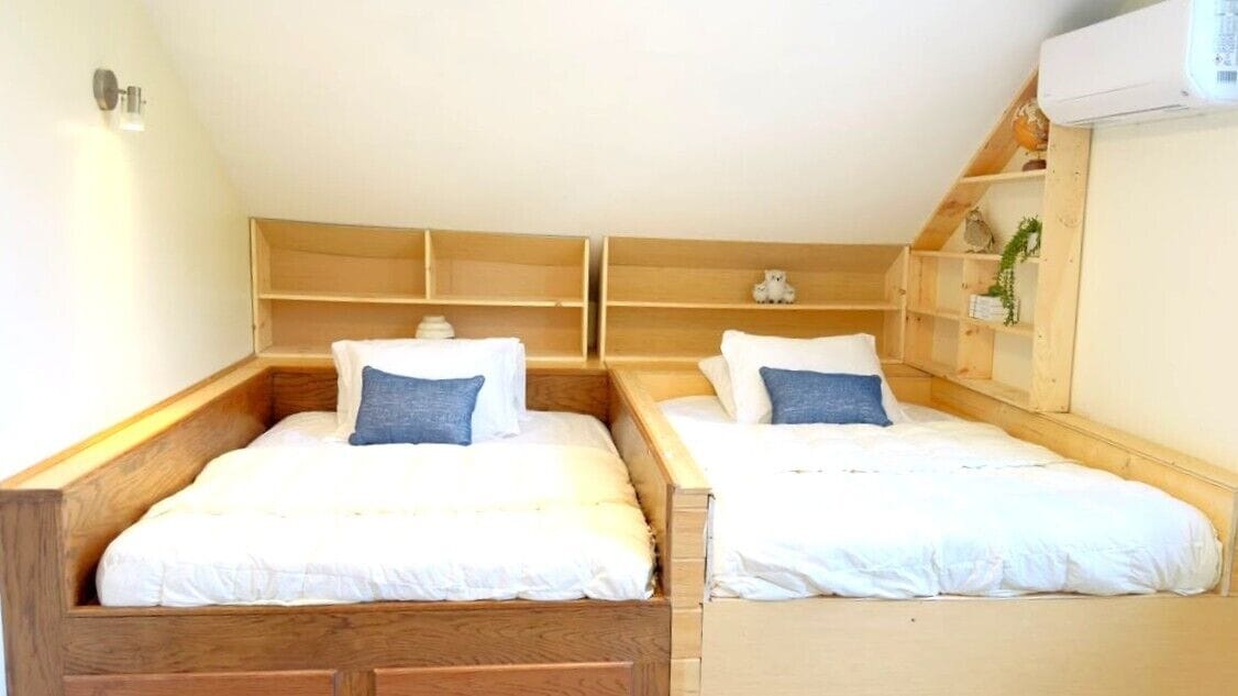 A room with two wooden beds, each with a white mattress and a blue pillow. There is a wooden shelf unit above the beds, and a small plant on the right side.