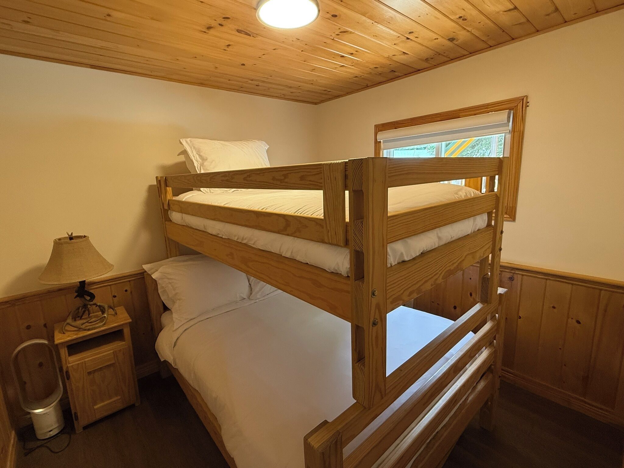 Habitación con literas, techo de madera, una ventana, una mesita de noche con lámpara y un armario pequeño.