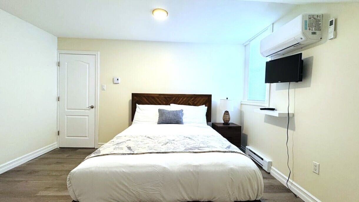 Une chambre à coucher comprenant un lit, une table de chevet, un téléviseur, un appareil de climatisation et une porte.