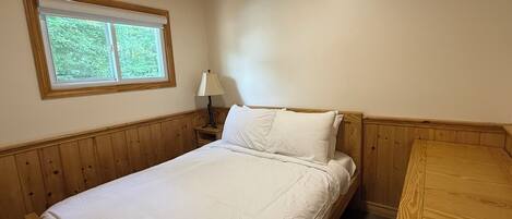 Un lit simple avec une tête de lit en bois, une table de chevet avec une lampe et une fenêtre donnant sur un paysage verdoyant.