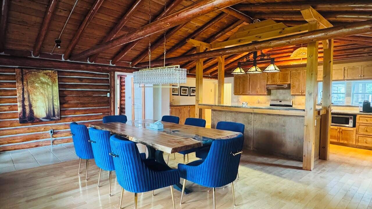 L’intérieur d’un chalet rustique est agrémenté d’une table à manger en bois, de chaises recouvertes d’un tissu bleu, d’un coin cuisine et d’un grand tableau accroché au mur.