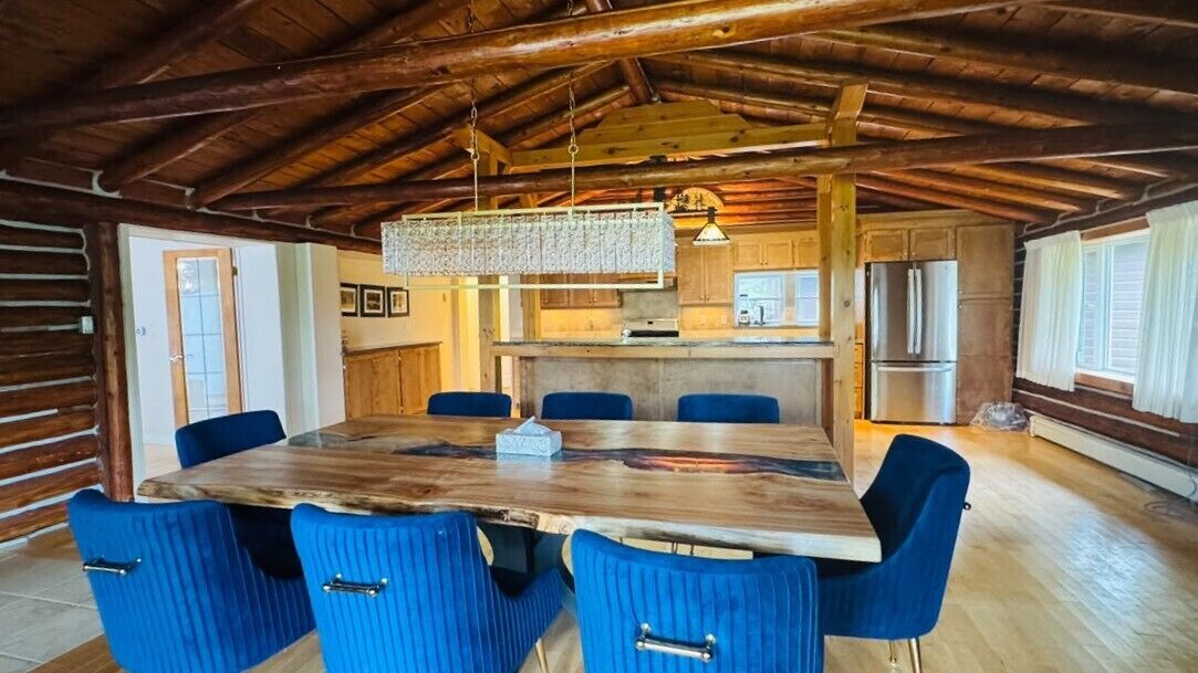 A rustic dining area with a large wooden table and blue upholstered chairs, a hanging light fixture, and a kitchen in the background.
