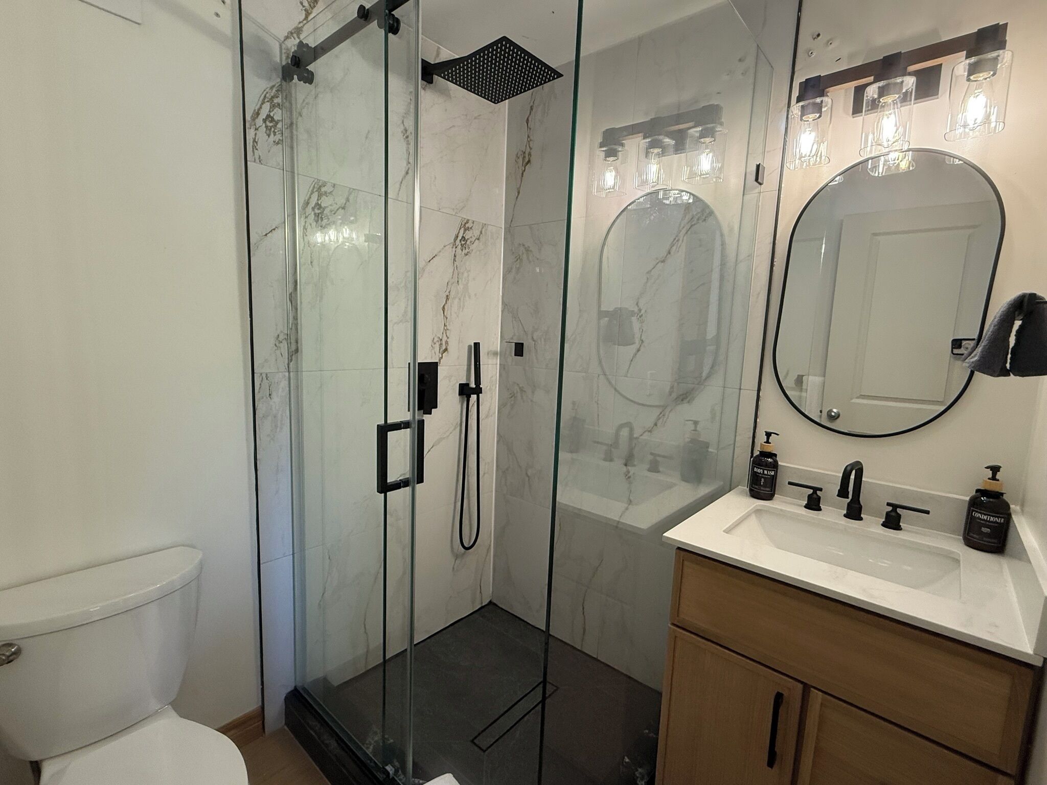 Un bany amb dutxa de vidre, vàter, un lavabo amb mirall i un moble de bany amb armaris.
