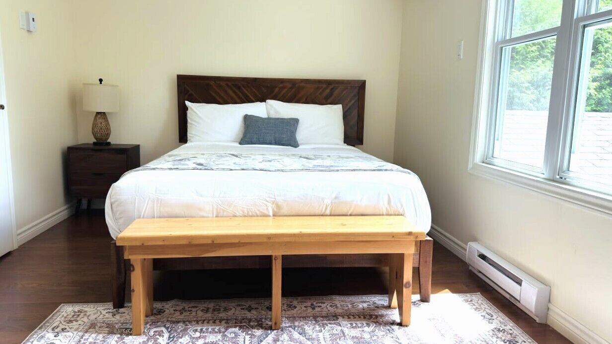 A bedroom with a bed, bedside table, lamp, bench, and a window.
