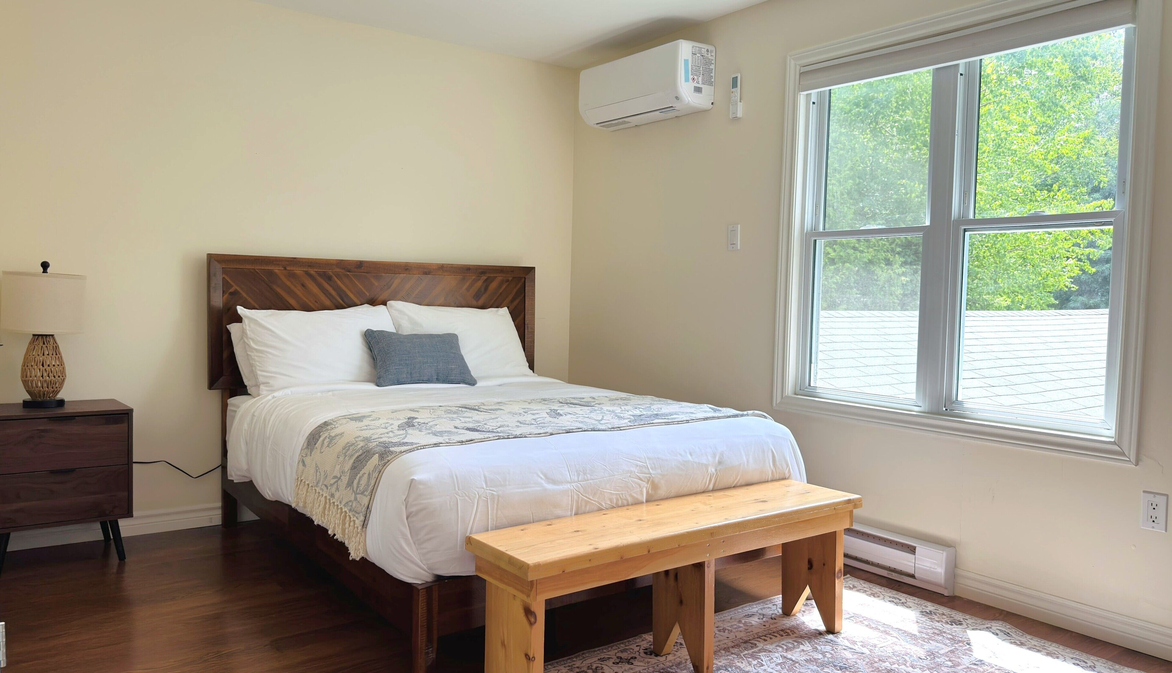 A bedroom with a bed, a bedside table, a lamp, a bench, and a window with a view of trees.