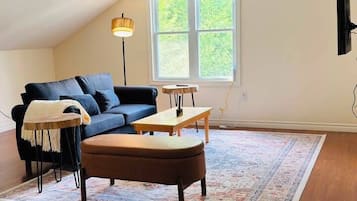 Um quarto estilo loft com um sofá azul escuro, uma mesa de centro de madeira, um tapete estampado e uma janela grande.