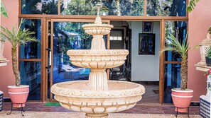 Une fontaine en pierre à plusieurs niveaux se trouve devant un bâtiment doté de portes vitrées, et de chaque côté, on peut voir des plantes en pot.