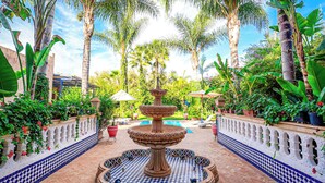 A courtyard with a central fountain, surrounded by tiled flooring and lush greenery.