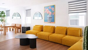 Een moderne woonkamer met een gele bank, een ronde salontafel en een schilderij van bloemen.