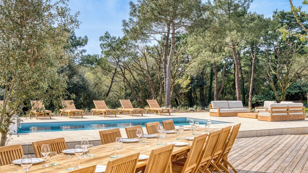Un área de comedor junto a la piscina con mesas y sillas de madera, piscina y sillones de descanso.