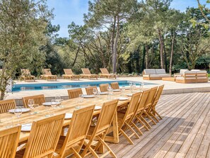 Un área de comedor junto a la piscina con mesas y sillas de madera, piscina y sillones de descanso.