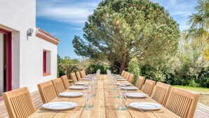 Una mesa de madera dispuesta para una comida, rodeada de sillas, con un edificio y árboles al fondo.