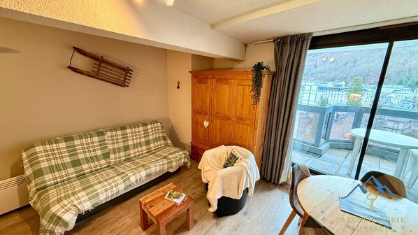 A cozy living room with a green plaid sofa, a wooden cabinet, a small table with a magazine, and a view of a cityscape through a large window. - Quiet Corner Near the Mountain Road (Mont-Dore)