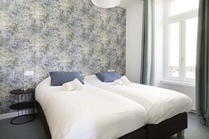 A double bed with white bedding and blue pillows, a small bedside table, and a floral wallpaper. - Villa Pax 3 - La Bourboule 6pers (La Bourboule)
