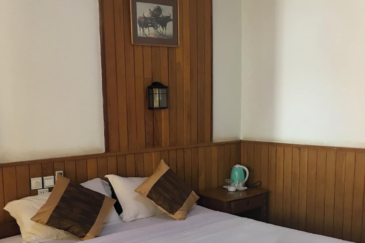 A hotel room with a wooden paneled headboard, a bed with white linens, two rolled towels, a nightstand with a teal lamp, and a framed picture on the wall.