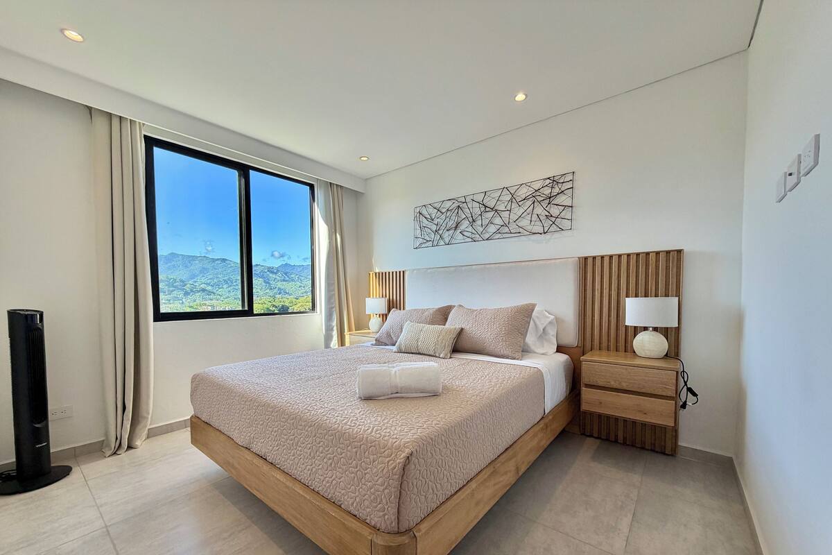 A modern hotel room with a large bed, a wooden headboard, bedside lamps, a flat-screen TV, and a view of mountains through the window.