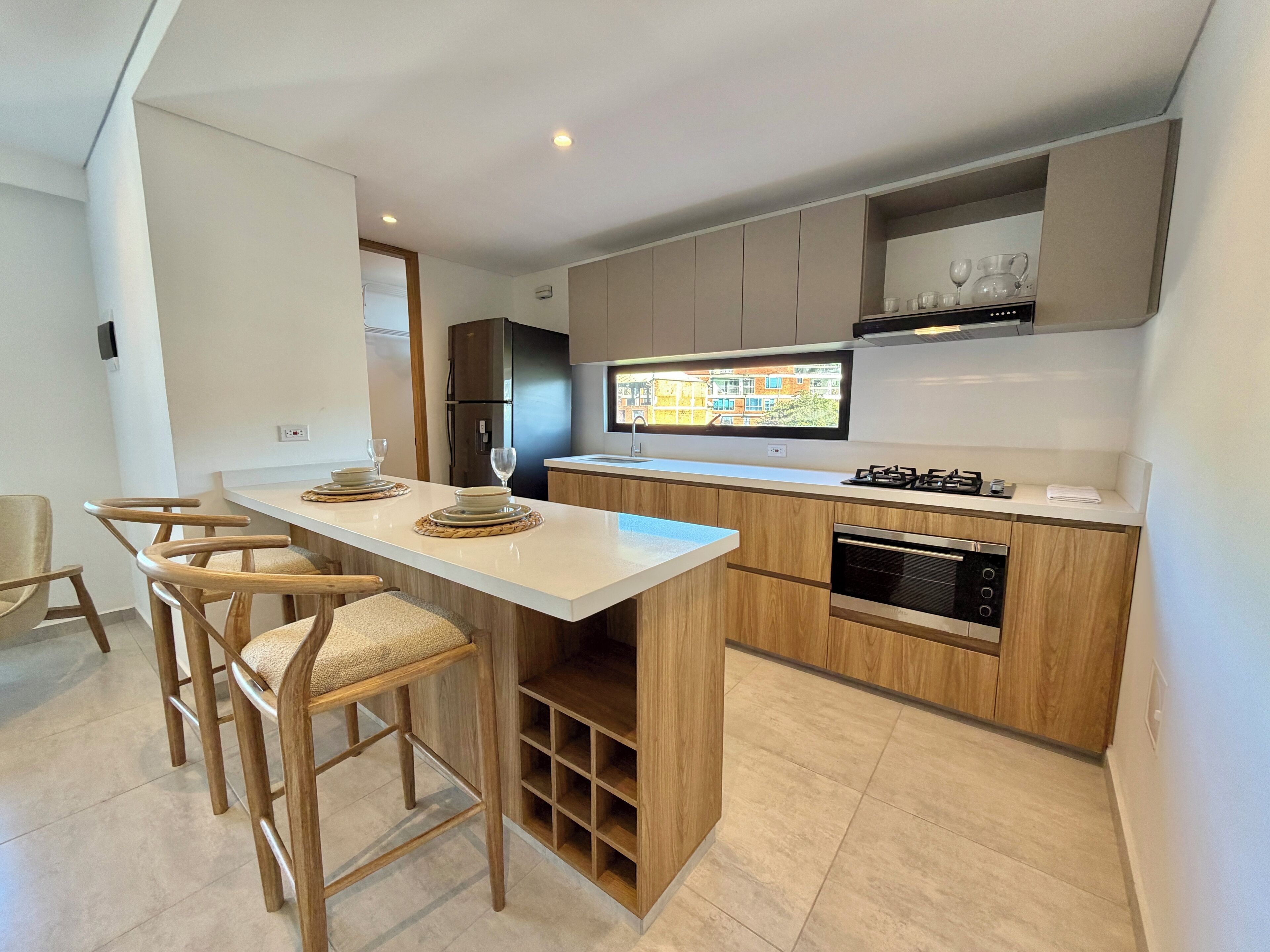 A modern kitchen with a central island, wooden chairs, a built-in oven, and a window overlooking a residential area.