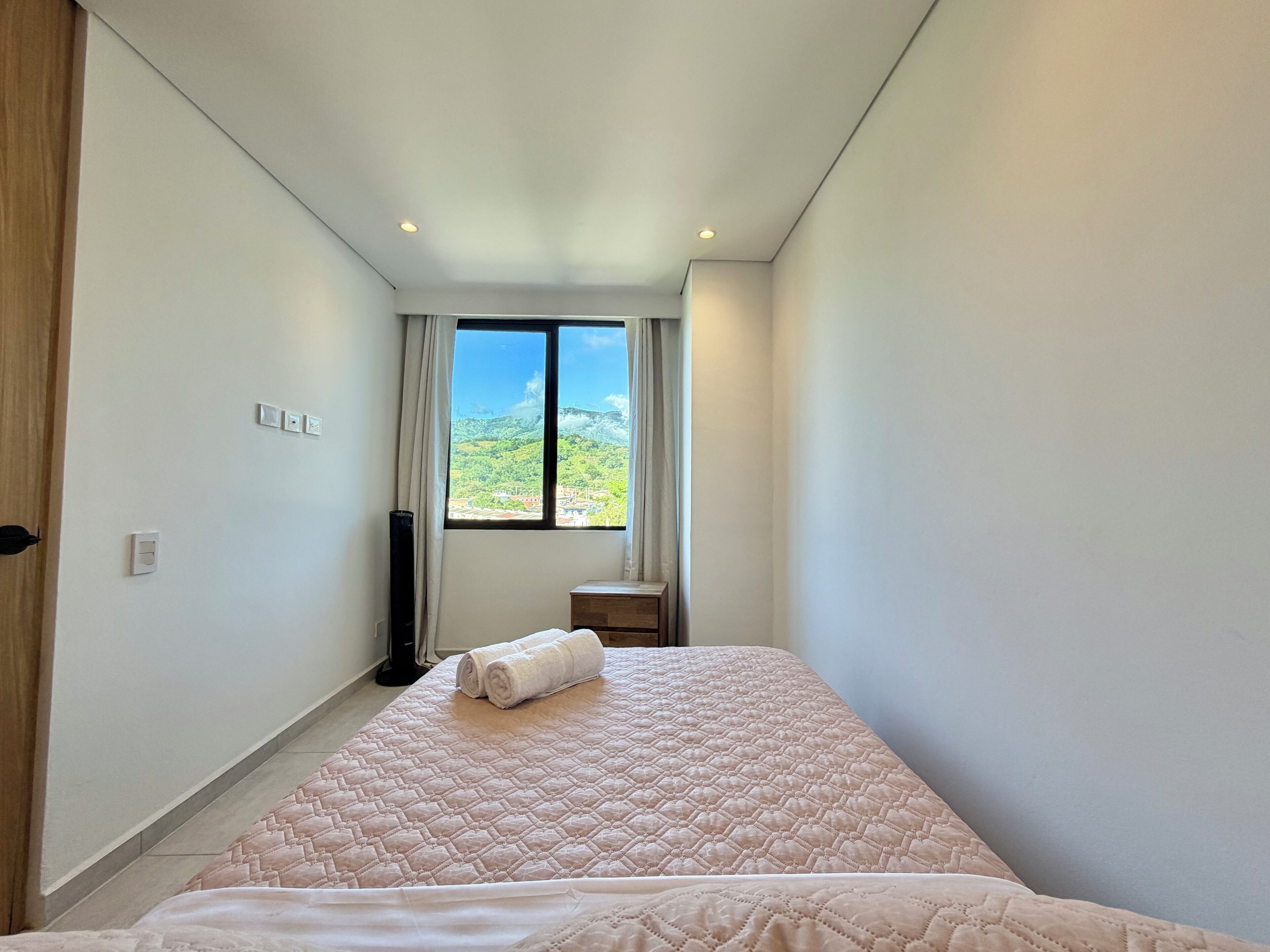 A hotel room with a large bed, a window with a view of a green landscape, a small bedside table, and a lamp.