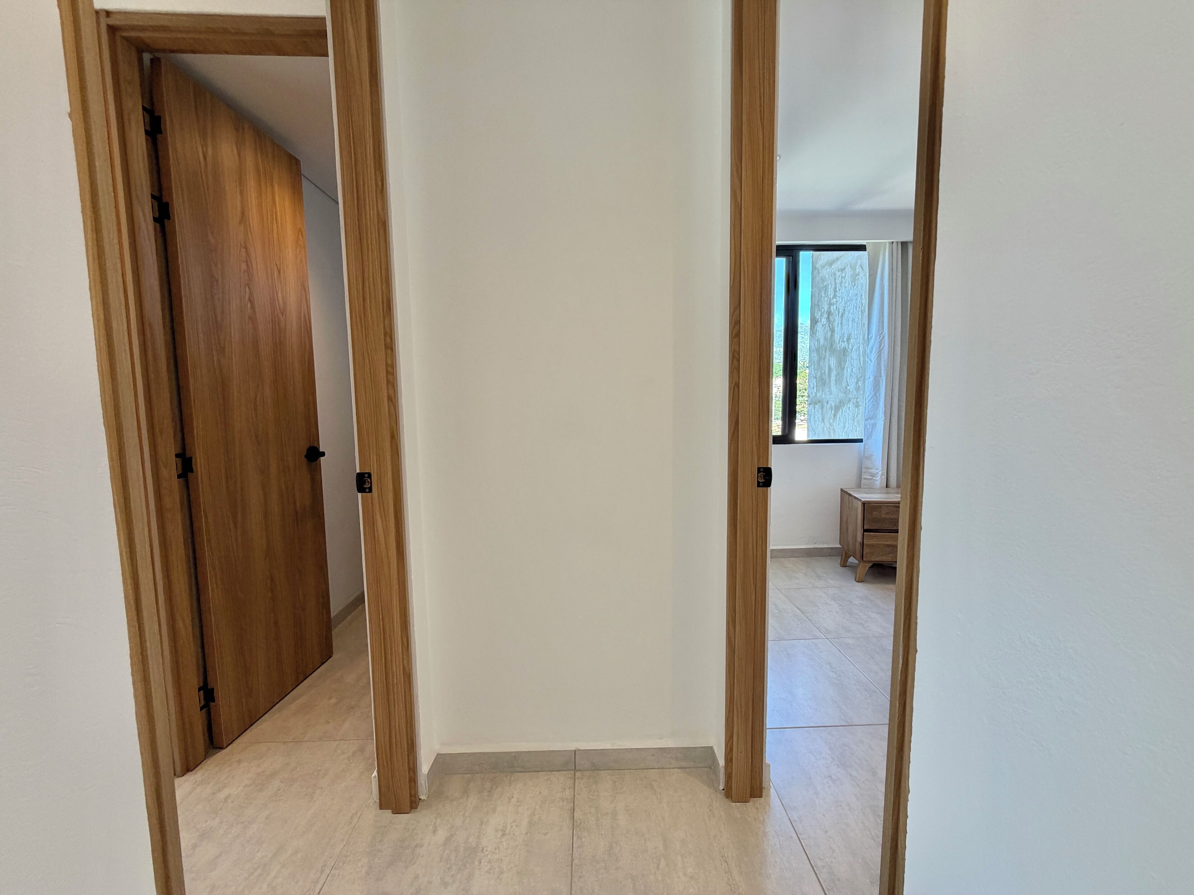 A hallway with wooden doors and a view of a room with a window and a small wooden table.