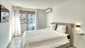 Quarto moderno com cama grande, janela com cortinas, ar condicionado fixado na parede e mesa de cabeceira com abajur.