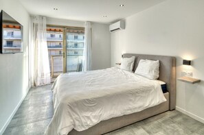 A modern bedroom with a large bed, a window with curtains, a wall-mounted air conditioner, and a bedside table with a lamp. - A/C 2bdr/6p Flat Just two Minutes From the Beach! (Antibes)