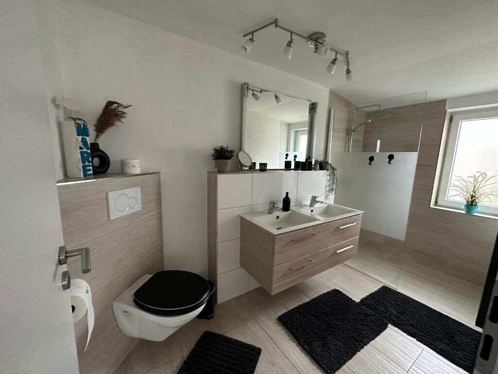 A modern bathroom with a toilet, sink, and mirror. There is a black rug on the floor and a window with a plant.