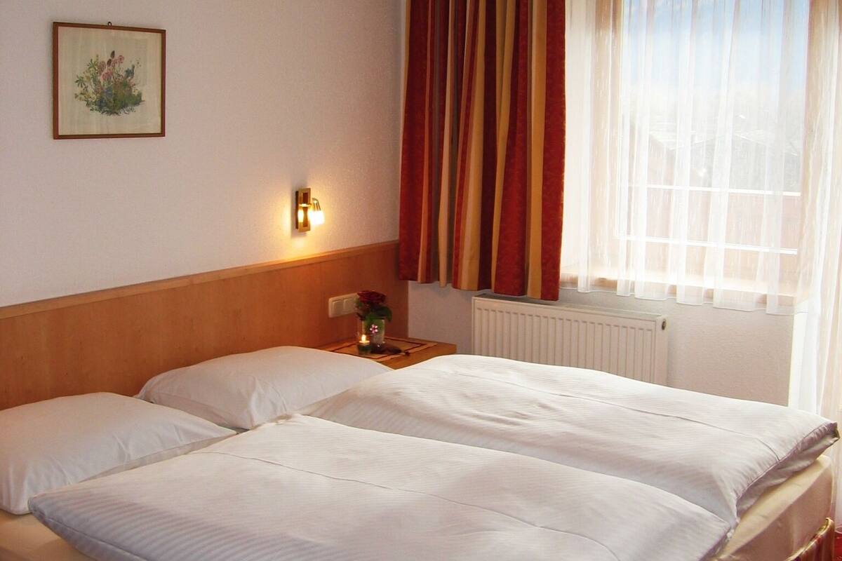 A double bed with white linens, a wooden headboard, a nightstand with a lamp, a framed picture on the wall, and a window with curtains.