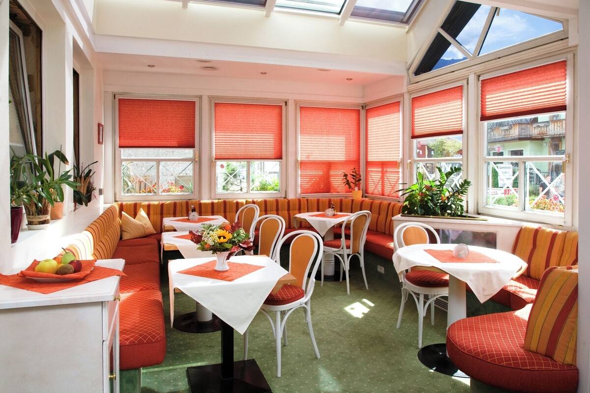 A sunlit dining area with a high ceiling, large windows with red blinds, and a variety of seating options including booths and chairs.