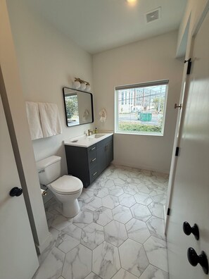Une salle de bain avec des toilettes, un lavabo et un miroir. Une fenêtre offre une vue sur l’extérieur.