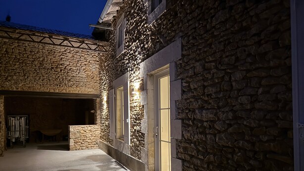 Um edifício de pedra com área externa coberta, uma porta visível e uma janela iluminada.