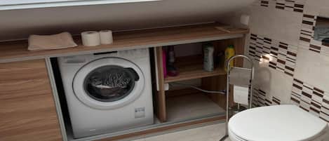 A compact bathroom with a built-in washing machine, a toilet, and a shelf with towels and toiletries.