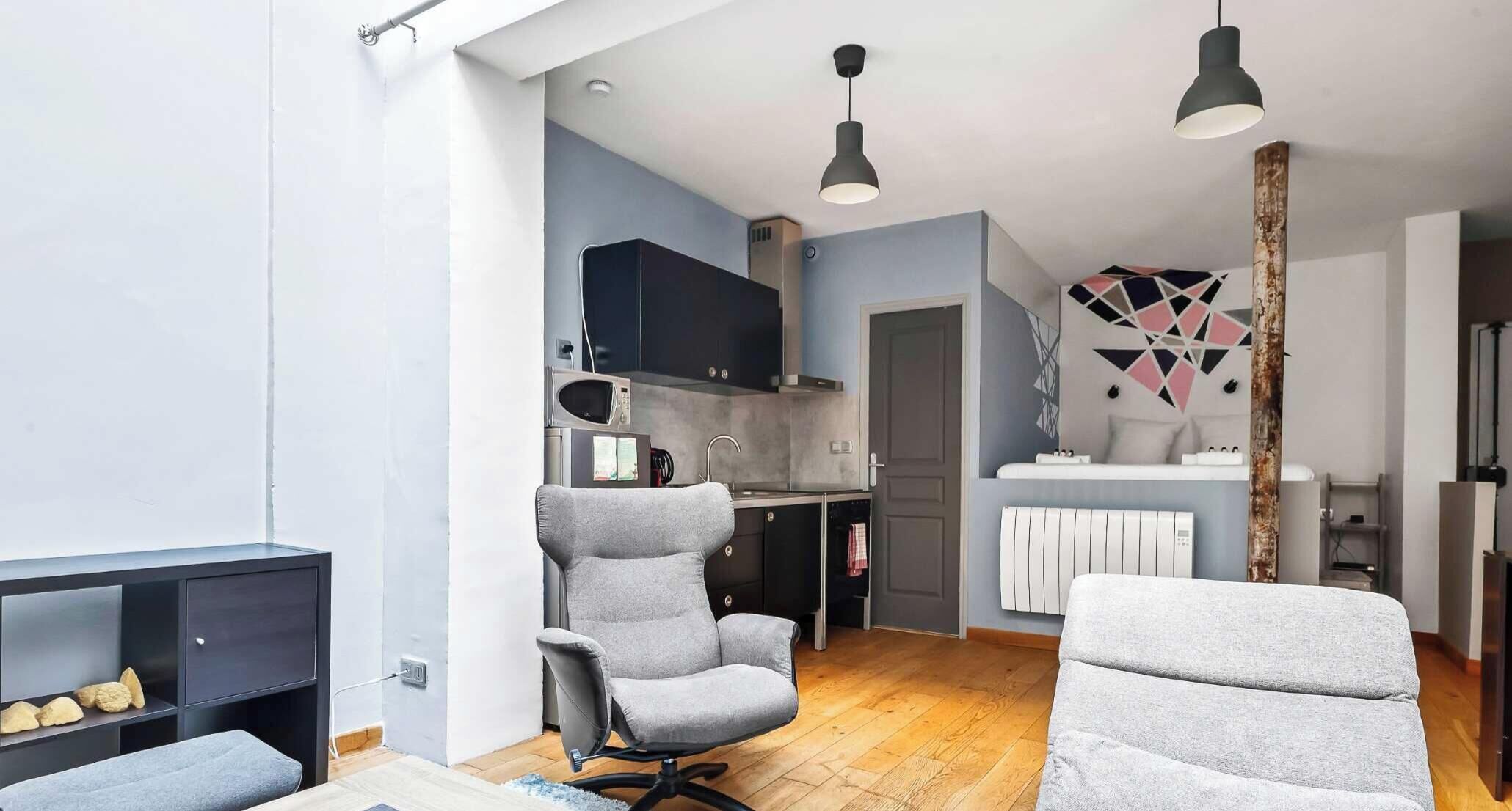 Un interior moderno con un sillón gris, un sofá, una zona de cocina con microondas y un elemento decorativo en la pared.