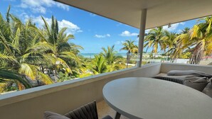 Ein Balkon mit rundem Tisch und gepolsterten Sitzgelegenheiten, der auf eine tropische Landschaft mit Palmen und Blick aufs Meer blickt.