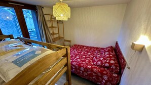 A room with a wooden bunk bed, a red floral bedspread, a ladder, a hanging light fixture, and a window with curtains. - Alpine Refuge Near the Forest Road (Mont-Dore)