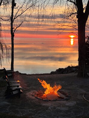 Sonnenuntergang über ruhigem Gewässer, im Vordergrund ein Lagerfeuer und Sitzgelegenheiten.
